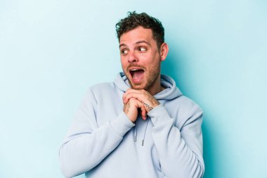 Kafkasyalı genç adam mavi arka planda izole edilmiş şans için dua ediyor, şaşırmış ve ağzını açıp önüne bakıyor..