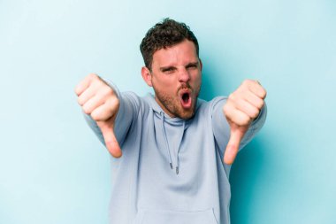 Mavi arka planda izole edilmiş genç beyaz bir adam baş parmağını aşağı gösteriyor ve hoşlanmadığını ifade ediyor..