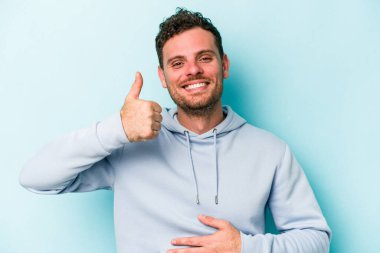 Mavi arka planda izole edilmiş genç beyaz adam karnına dokunuyor, nazikçe gülümsüyor, yemek yiyor ve tatmin oluyor..