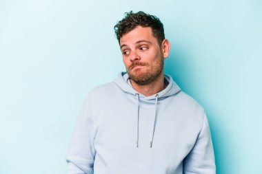 Mavi arka planda izole edilmiş genç beyaz adam omuzlarını silkeliyor ve gözleri açık..