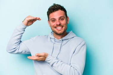 Kafkasyalı genç adam mavi arka planda izole edilmiş. Elinde işaret parmaklarıyla küçük bir şey tutuyor, gülümsüyor ve kendinden emin..