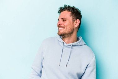 Kafkasyalı genç adam mavi arka planda izole edilmiş rahat ve mutlu bir şekilde gülüyor, boynunda dişlerini gösteriyor..