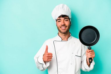 Kafkasyalı genç aşçı elinde uçan tavayla mavi arka planda izole edilmiş gülümsüyor ve başparmağını kaldırıyor.