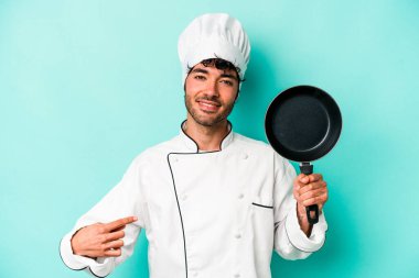 Kafkasyalı genç aşçı elinde uçan tavayla mavi arka planda izole edilmiş gömlek fotokopi alanını işaret eden gururlu ve kendinden emin.