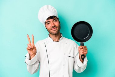 Kafkasyalı genç aşçı elinde uçan tavayla mavi arka planda iki numarayı parmaklarıyla gösteriyor..