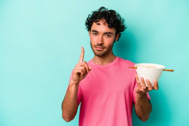 Mavi arka planda izole edilmiş erişte yiyen genç beyaz adam parmak ile bir numarayı gösteriyor..