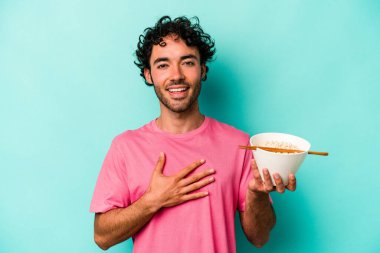 Mavi arka planda izole edilmiş erişte yiyen genç beyaz adam yüksek sesle gülüyor ve elini göğsüne koyuyor..