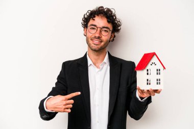 Beyaz arka planda izole edilmiş oyuncak evi tutan beyaz tenli genç beyaz iş adamı gömlek fotokopi alanını işaret ediyor, gururlu ve kendinden emin.