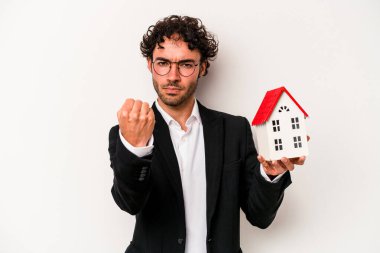 Beyaz arka planda izole edilmiş oyuncak evi tutan Kafkasyalı genç bir iş adamı kameraya yumruk gösteriyor, agresif bir yüz ifadesi..