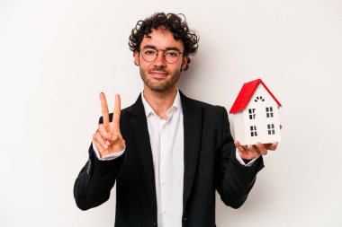 Beyaz arka planda iki numarayı parmaklarıyla gösteren bir oyuncak evi tutan Kafkasyalı genç iş adamı..