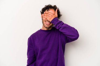 Beyaz arka planda soyutlanmış beyaz tenli genç bir adam elleriyle gözlerini kapatıyor, geniş bir gülümsemeyle bir sürpriz bekliyor..