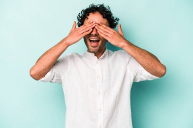 Beyaz arka planda soyutlanmış beyaz tenli genç bir adam elleriyle gözlerini kapatıyor, geniş bir gülümsemeyle bir sürpriz bekliyor..