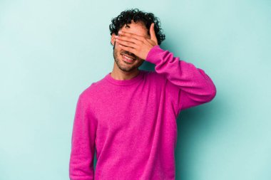 Beyaz arka planda soyutlanmış beyaz tenli genç bir adam elleriyle gözlerini kapatıyor, geniş bir gülümsemeyle bir sürpriz bekliyor..