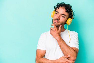 Mavi arka planda izole edilmiş müzik dinleyen beyaz bir adam şüpheli ve şüpheci bir ifadeyle yana bakıyor..