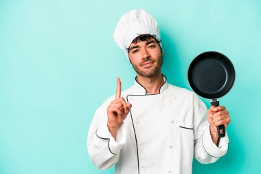 Kafkasyalı genç aşçı elinde uçan bir tavayla mavi arka planda izole edilmiş. Bir numarayı parmağıyla gösteriyor..