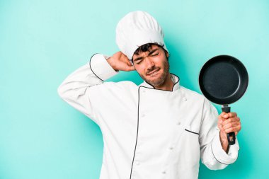 Kafkasyalı genç aşçı elinde uçan tavayla mavi arka planda izole edilmiş kafasının arkasına dokunuyor, düşünüyor ve bir seçim yapıyor..