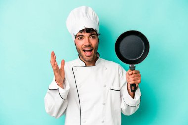 Kafkasyalı genç aşçı elinde uçan bir tavayla mavi arka planda izole edilmiş güzel bir sürpriz bekliyor, heyecanlı ve el kaldırıyor..