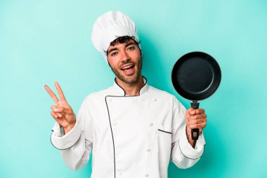 Kafkasyalı genç aşçı elinde uçan tavayla mavi arka planda izole edilmiş neşeli ve tasasız parmaklarıyla barış sembolü gösteriyor..