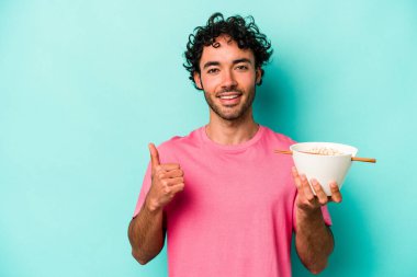 Mavi arka planda izole edilmiş erişte yiyen genç beyaz adam gülümsüyor ve başparmağını kaldırıyor.