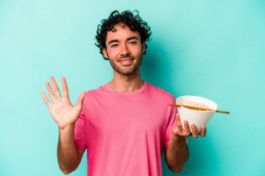 Mavi arka planda izole edilmiş erişte yiyen genç beyaz adam gülümseyerek beş numarayı parmaklarıyla gösteriyor..