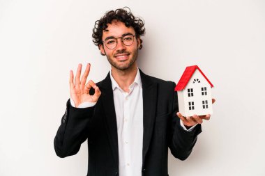 Beyaz arka planda izole edilmiş oyuncak evi tutan Kafkasyalı genç bir iş adamı neşeli ve kendinden emin bir şekilde iyi niyet gösteriyor..