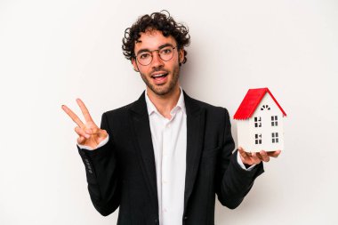 Beyaz arka planda izole edilmiş bir oyuncak evi tutan Kafkasyalı genç bir iş adamı neşeli ve kaygısız parmakları olan bir barış sembolü gösteriyor..