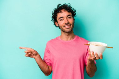 Mavi arka planda izole edilmiş erişte yiyen genç beyaz adam gülümsüyor ve boş alanda bir şeyler gösteriyor..