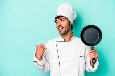 Kafkasyalı genç aşçı elinde uçan tavayla mavi arka planda izole edilmiş, başparmak uzakta, gülerek ve kaygısızca.
