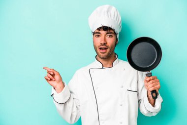 Kafkasyalı genç aşçı elinde uçan bir tava tutuyor. Mavi arka planda, yan tarafı işaret ediyor.