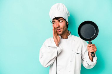 Kafkasyalı genç aşçı elinde uçan bir tavayla mavi arka planda izole edilmiş bir şekilde gizli bir sıcak fren haberi veriyor ve yana bakıyor.