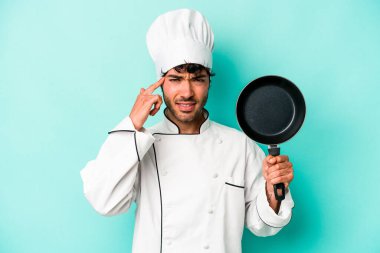 Kafkasyalı genç aşçı elinde uçan tavayla mavi arka planda izole edilmiş işaret parmağıyla hayal kırıklığı jesti yapıyor..