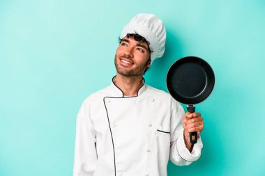 Kafkasyalı genç aşçı elinde uçan tavayla mavi arka planda tek başına hedeflere ve amaçlara ulaşmayı hayal ediyor.