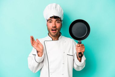 Kafkasyalı genç aşçı elinde uçan tavayla mavi arka planda izole edilmiş. Şaşırmış ve şok olmuş..