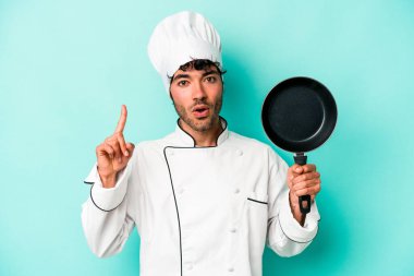 Kafkasyalı genç aşçı elinde uçan tavayla mavi arka planda izole edilmiş bir şekilde bir fikri, ilham verici bir fikri var..