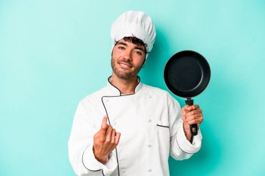 Kafkasyalı genç aşçı elinde uçan tavayla mavi arka planda izole edilmiş bir şekilde seni işaret ediyor sanki davet ediyormuş gibi..