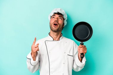 Kafkasyalı genç aşçı elinde uçan tavayla mavi arka planda izole edilmiş ağzı açık bir şekilde yukarı işaret ediyor..