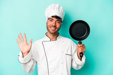 Kafkasyalı genç aşçı elinde uçan tavayla mavi arka planda izole edilmiş gülümseyen beş numarayı parmaklarıyla gösteriyor..