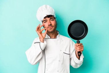 Kafkasyalı genç aşçı elinde uçan tavayla mavi arka planda izole edilmiş dudaklarında parmaklarıyla bir sır saklıyor..