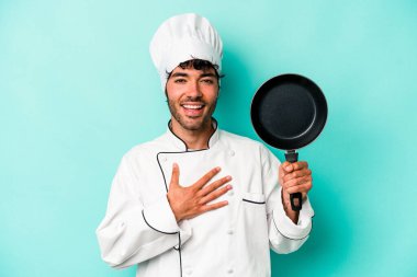 Kafkasyalı genç aşçı elinde uçan bir tavayla mavi arka planda izole bir şekilde gülüyor ve elini göğsünde tutuyor..