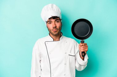 Kafkasyalı genç aşçı elinde uçan tavayla mavi arka planda izole edilmiş omuzlarını silker ve gözleri açık kalır..