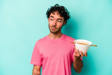 Mavi arka planda izole edilmiş erişte yiyen genç beyaz adam şaşkın ve kararsız hissediyor..