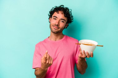 Mavi arka planda izole edilmiş erişte yiyen genç beyaz adam sanki seni davet ediyormuş gibi seni işaret ediyor..