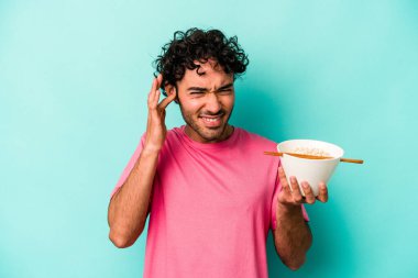 Mavi arka planda izole edilmiş erişte yiyen genç beyaz adam elleriyle kulaklarını kapatıyor..