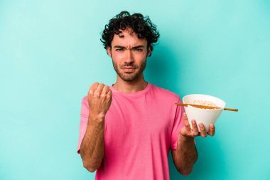 Mavi arka planda izole edilmiş erişte yiyen genç beyaz adam kameraya yumruk gösteriyor, agresif yüz ifadesi var..