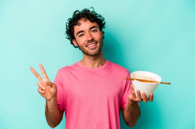 Mavi arka planda izole edilmiş erişte yiyen genç beyaz adam neşeli ve kaygısız parmaklarıyla barış sembolü gösteriyor..