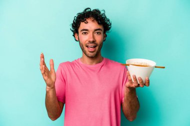 Mavi arka planda izole edilmiş erişte yiyen genç beyaz adam hoş bir sürpriz alıyor, heyecanlı ve el kaldırıyor..