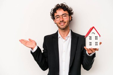 Beyaz arka planda izole edilmiş oyuncak evi tutan Kafkasyalı genç bir iş adamı bir avuç içinde fotokopi alanı ve belinde bir el daha gösteriyor..