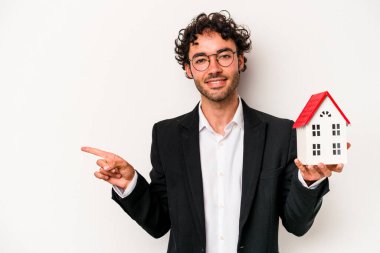 Beyaz arka planda izole edilmiş bir oyuncak evi tutan beyaz iş adamı gülümsüyor ve boş alanda bir şey gösteriyor..