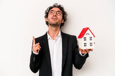 Beyaz arka planda izole edilmiş bir oyuncak evi tutan Kafkasyalı genç iş adamı ağzı açık bir şekilde üst tarafı işaret ediyor..