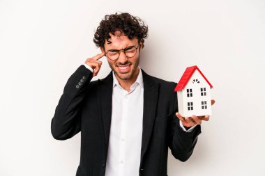 Beyaz arka planda izole edilmiş bir oyuncak evi tutan Kafkasyalı genç bir iş adamı elleriyle kulaklarını kapatıyor..